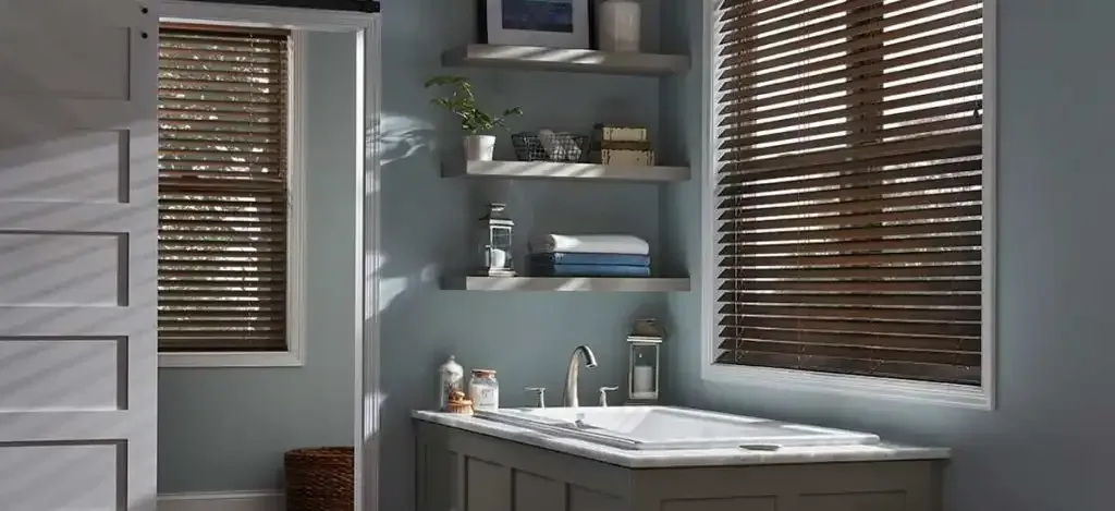 Bathroom with a bathtub, shelves, and window with blinds. Soft blue walls and natural light.
