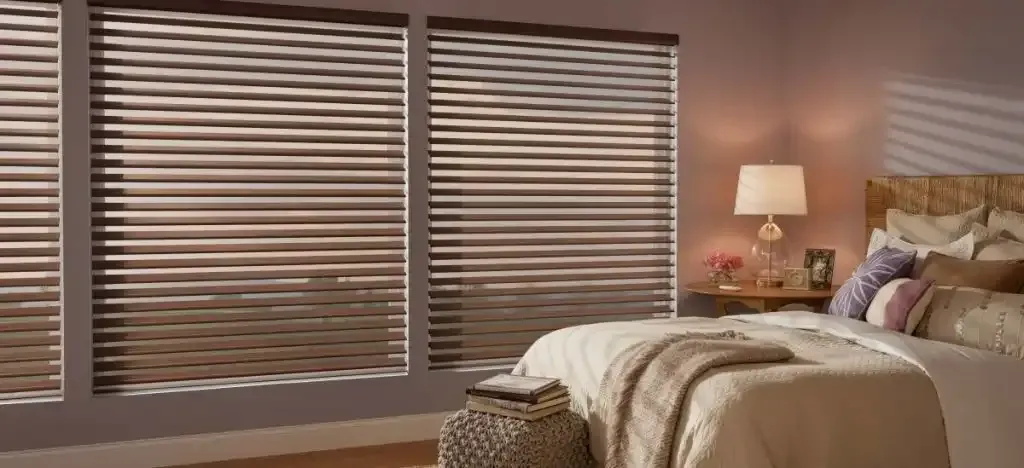 Bedroom with closed horizontal blinds and a bed with pillows and a lamp on a nightstand.