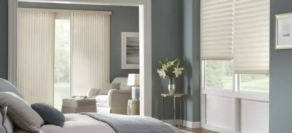 Bedroom with blue walls, window blinds, curtains, and a bed. A side table has flowers.