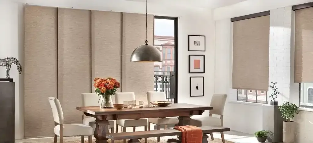 Dining room with large wood table, upholstered chairs, fabric wall panels, and window shades.